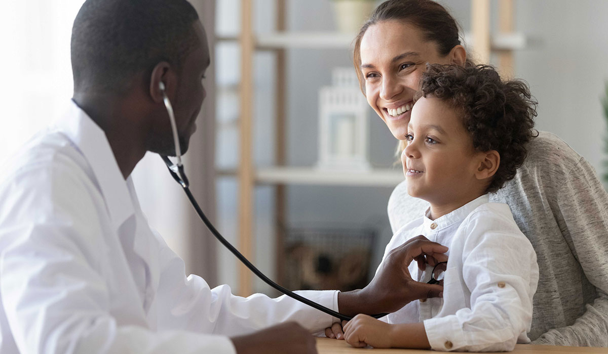 A Physician listening to a smiling little boy's heart with a stethoscope while a smiling mother looks on.